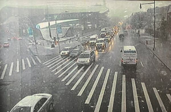 Grey city intersection with zebra crossings, traffic, and a bus moving through rain or haze.